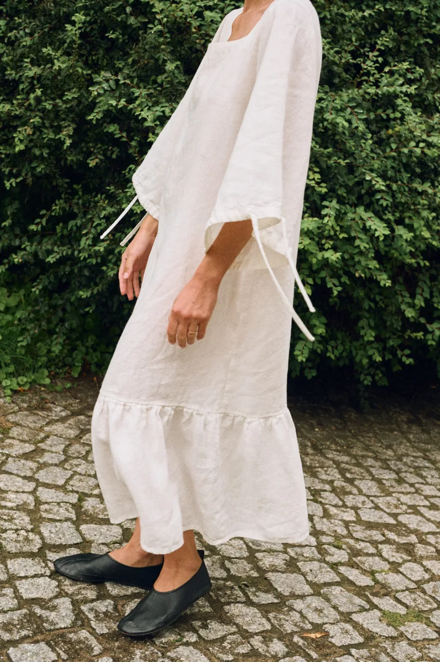 Model wearing an off-white linen midi dress with wide sleeves and ruffle hem, side view highlighting the natural linen drape