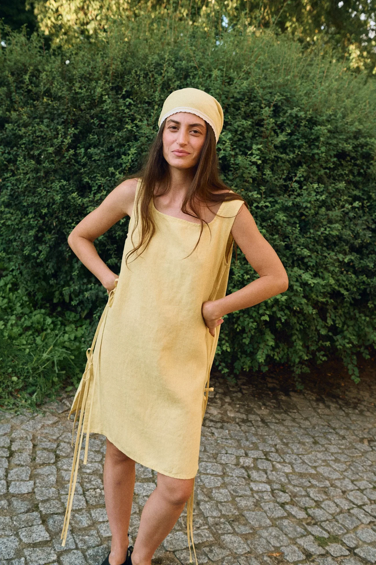 Model wearing a sleeveless butter yellow linen midi dress with a scoop neckline, side tie details, and a matching linen head wrap bandana