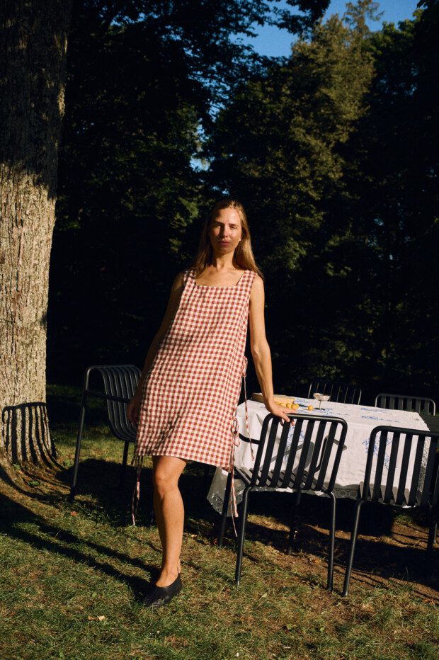 Woman wearing a sleeveless dress in terracotta gingham linen, featuring a square neckline, bust darts for gentle bust shaping, side ties, and a relaxed, breathable silhouette with a midi-length hem