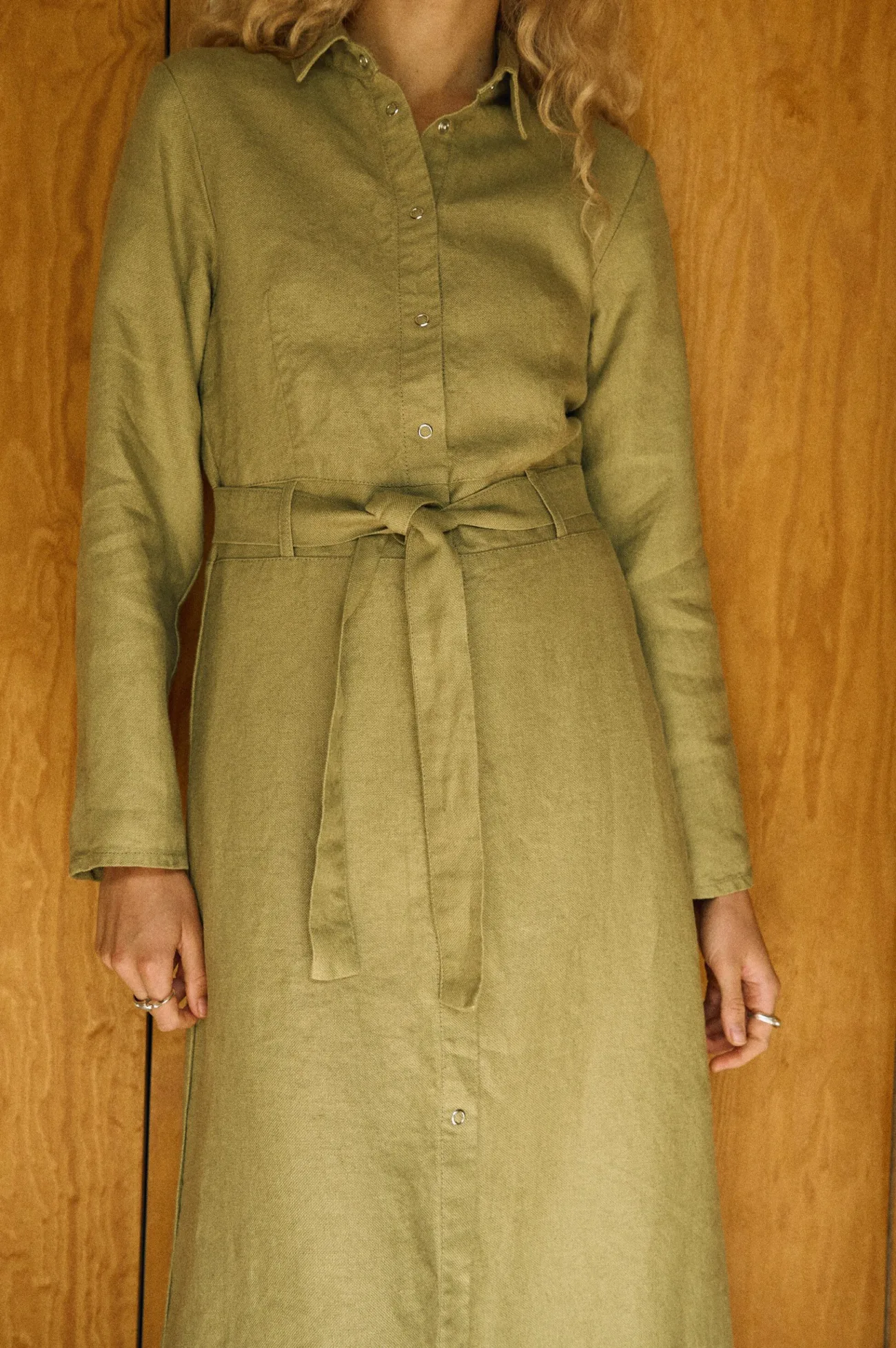 Close-up of an olive heavy linen shirt dress featuring a snap-front closure, long sleeves, and a matching belt that defines the waist