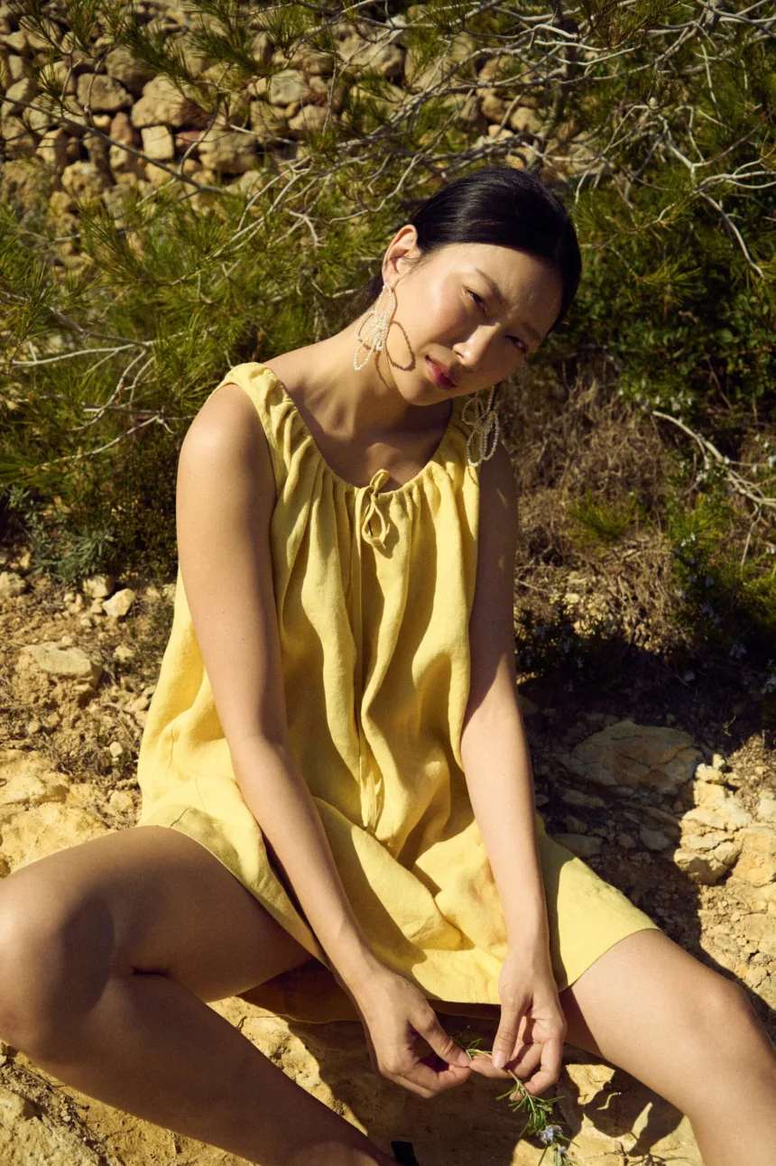 Sleeveless butter yellow linen mini dress with a gathered tie neckline and loose above-knee silhouette, shown in a seated front view