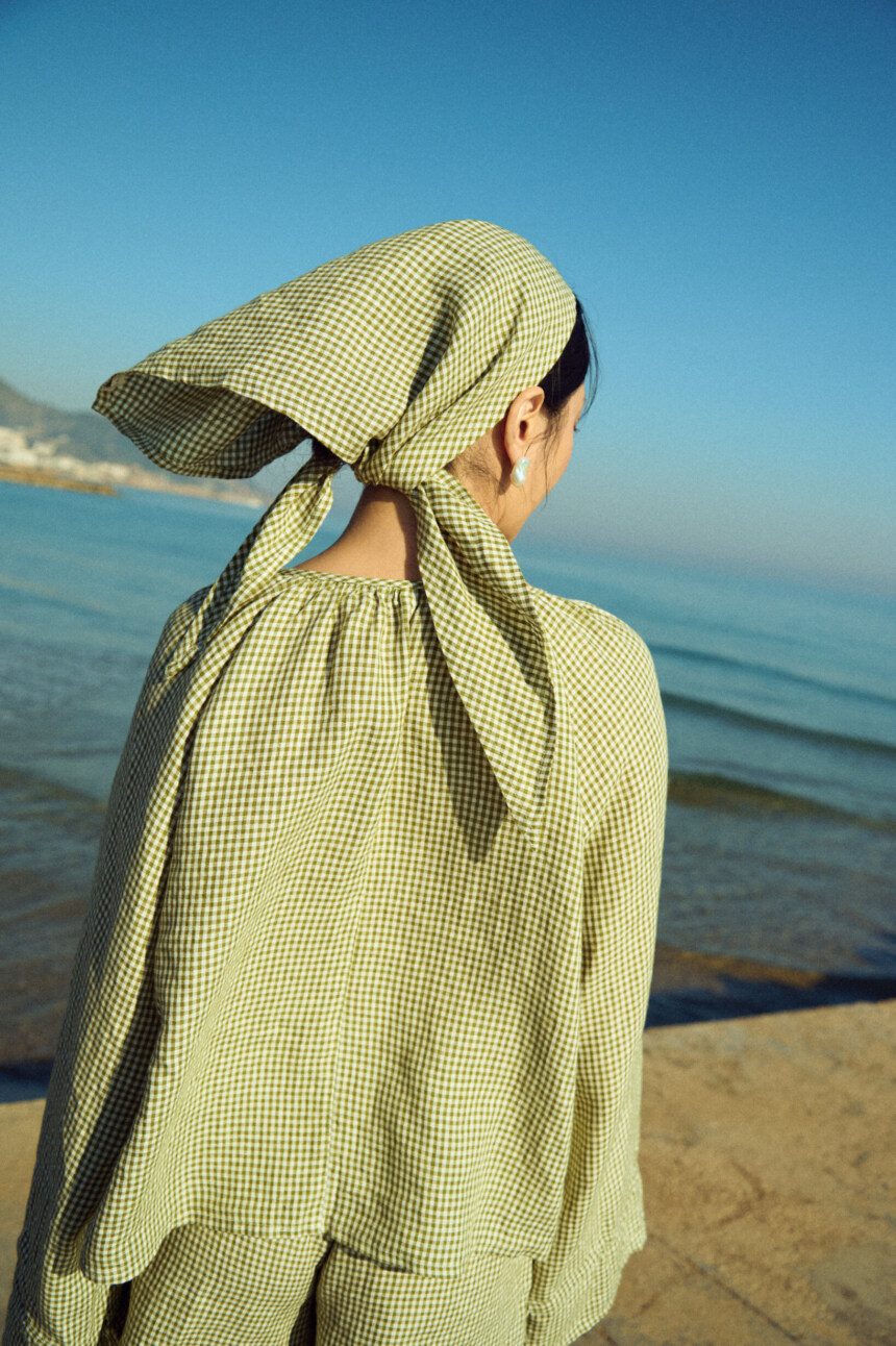 Back view of a moss green mini gingham linen triangle scarf tied at the nape, worn with a matching airy gingham blouse
