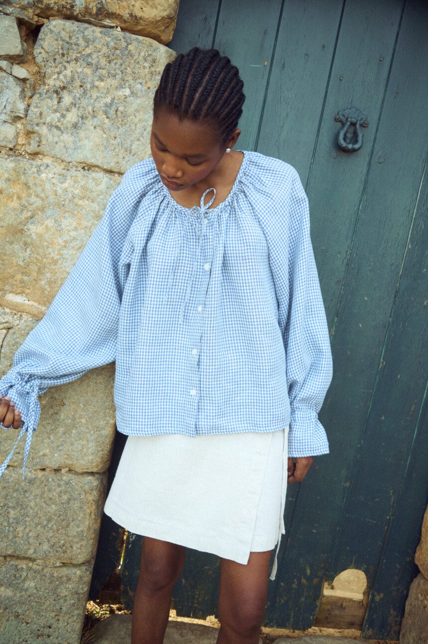 Blue gingham linen blouse with soft gathers at the neckline and long sleeves with tie details, worn with a light linen mini skirt