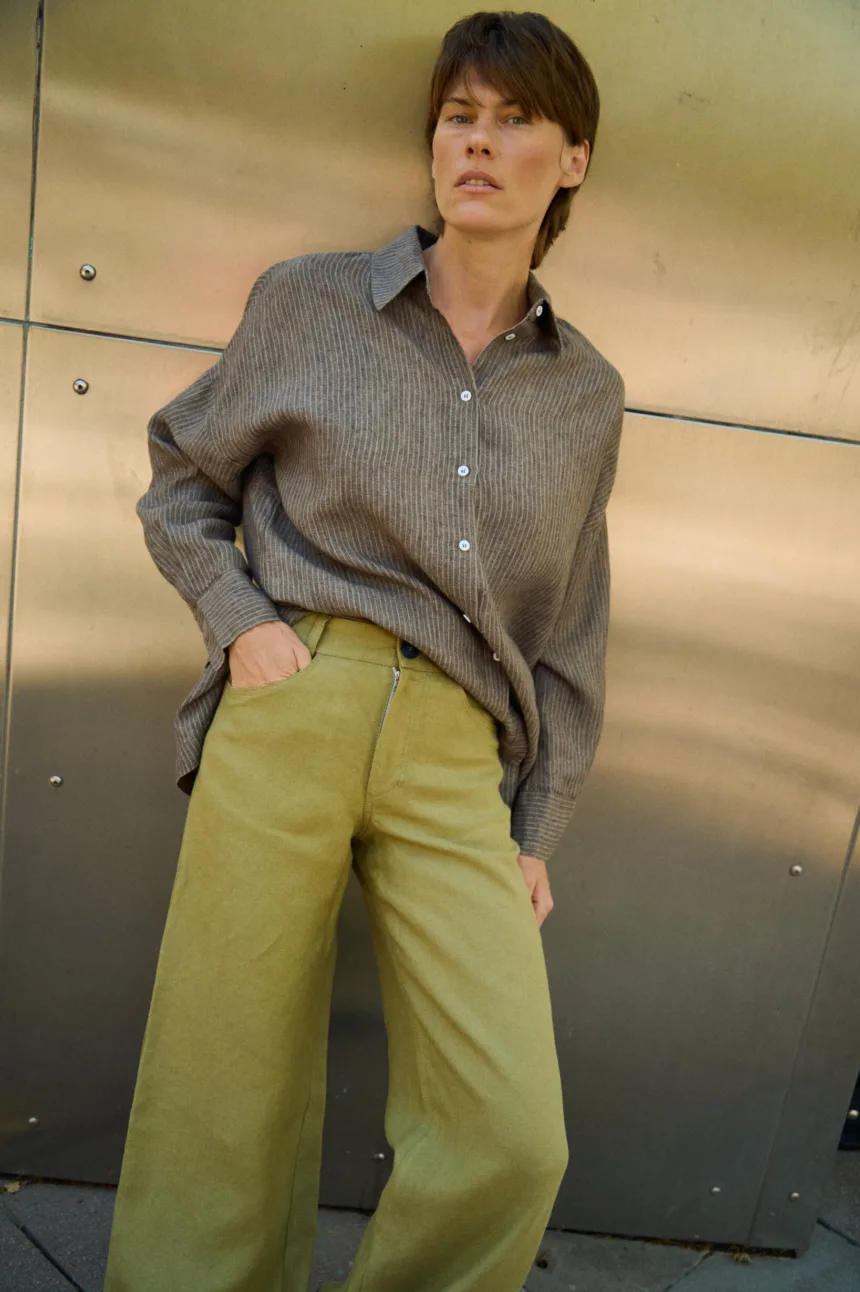 Close-up of olive heavy linen pants showing the front button closure and curved side pocket, styled with a brown striped linen shirt