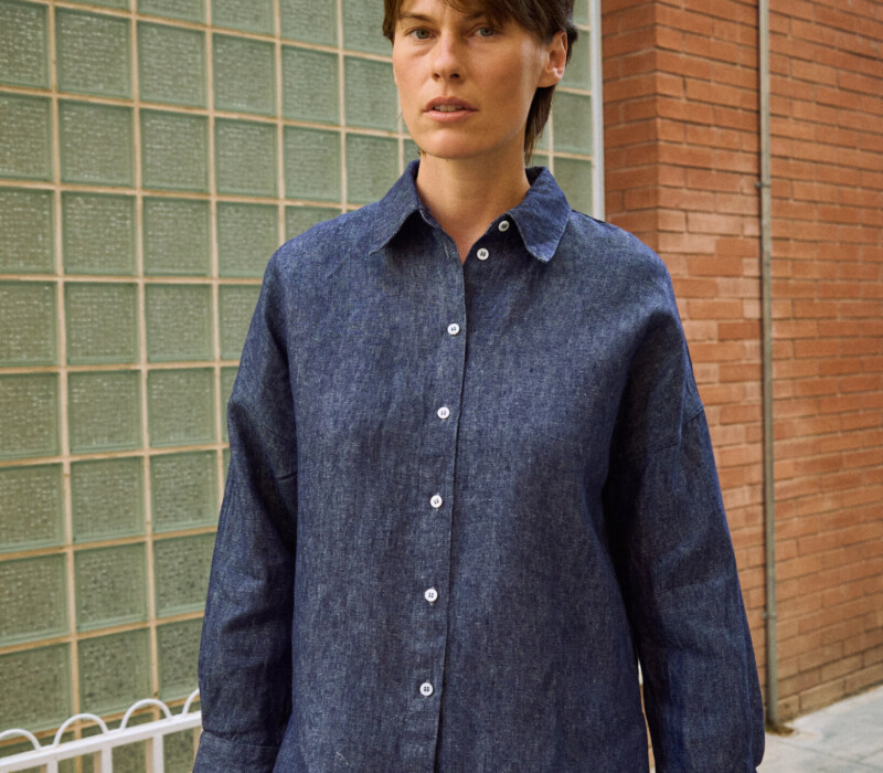 Front detail of blue denim linen shirt with button-down front, pointed collar, dropped shoulders, and wide cuffs