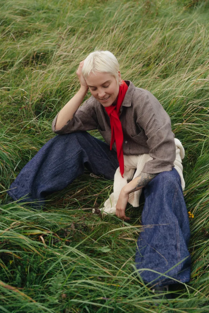 Brown striped linen shirt styled with a bright red neck scarf and cream knit sweater, worn with wide-leg denim linen trousers